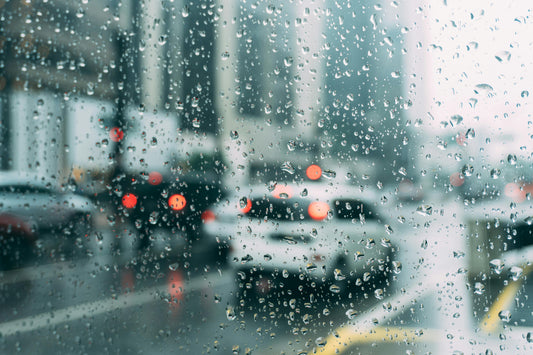Photo of window in car on Rainy day with water droplets on the window, showing being in traffic in a city.